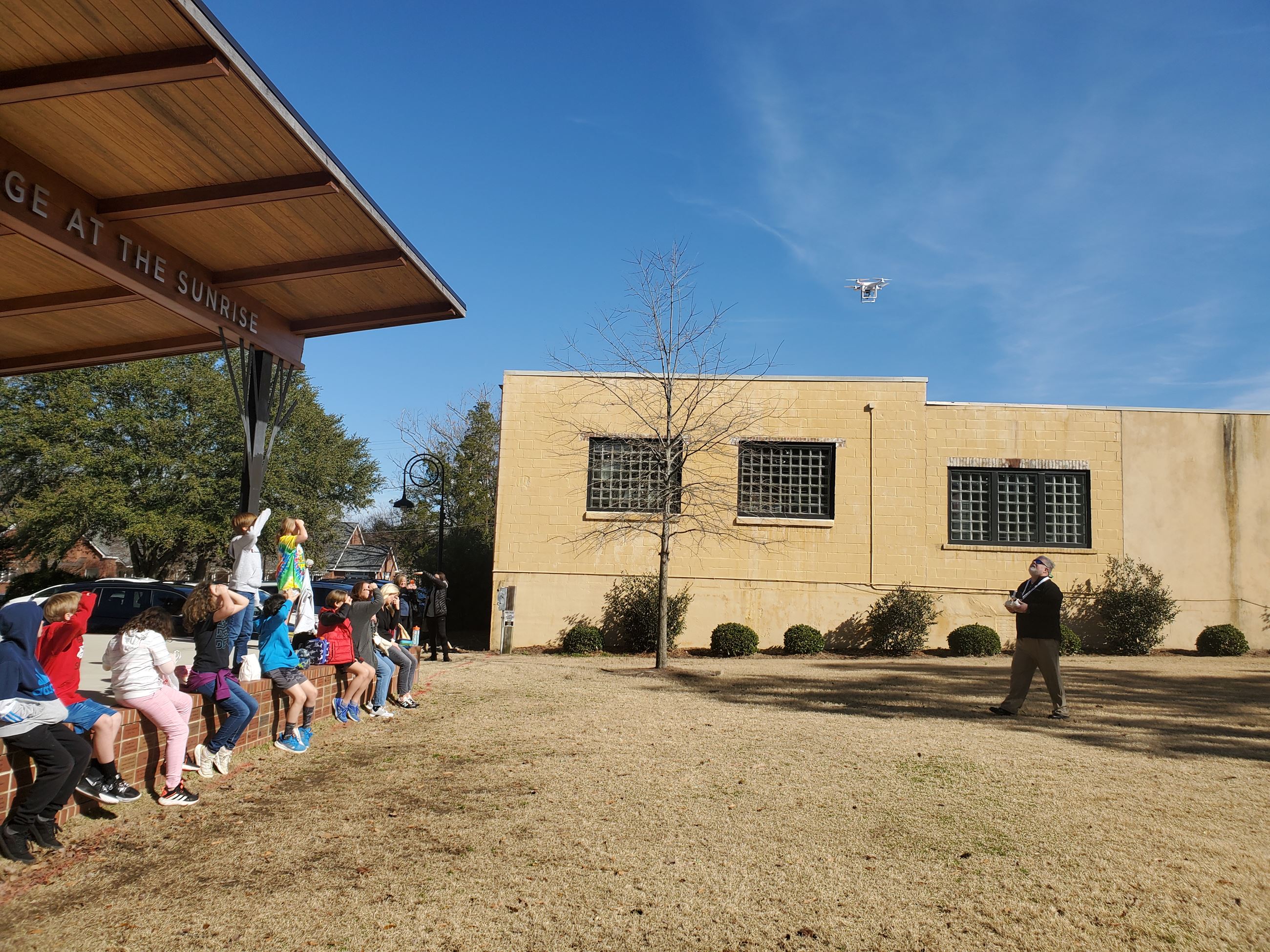 School children watch a man fly a drone in a grassy area